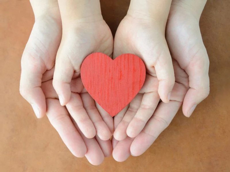 A mother's hands holding a child's hands which hold a heart.
