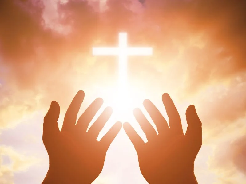 Hands of a saint raised in prayer with a cross in the background